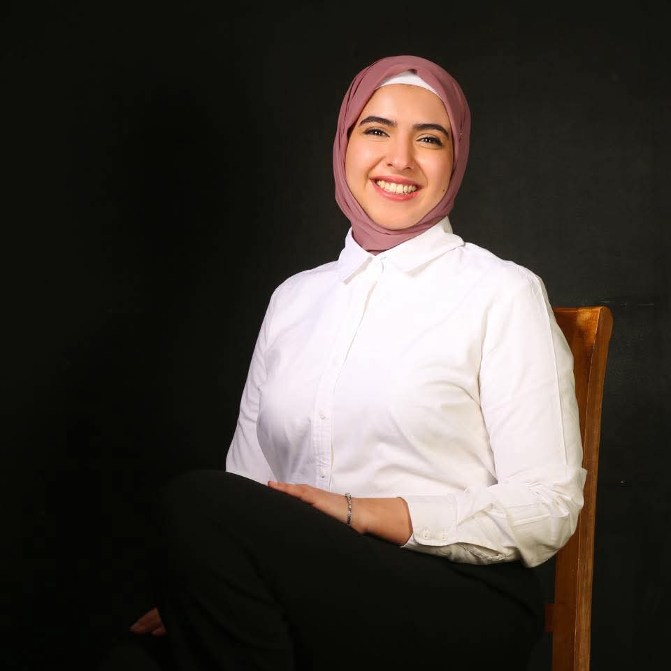 Alaa El Emam seated, wearing a white shirt and a pink hijab